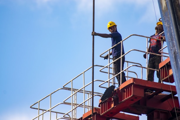 Two construction workers stand on a red metal scaffolding structure. They are wearing blue shirts and yellow safety helmets, suggesting an environment focused on safety and construction. One worker is holding onto a long metal rod as they both look into the distance under a clear blue sky.