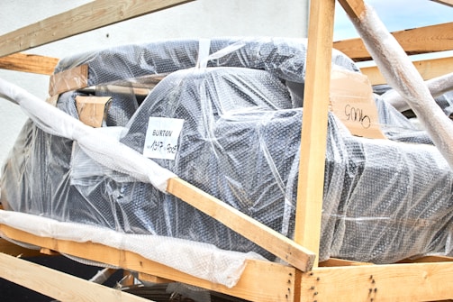 A large object wrapped in bubble wrap is secured within a wooden frame. There are some labels and markings on the packaging, with one label reading 'BURTON'. The object appears to be prepared for shipping or transport.