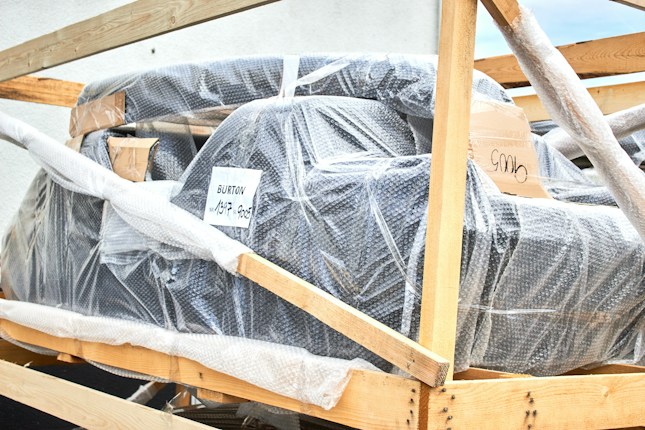 A large object wrapped in bubble wrap is secured within a wooden frame. There are some labels and markings on the packaging, with one label reading 'BURTON'. The object appears to be prepared for shipping or transport.