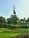 A tall, conical tree is positioned at the center of a well-maintained circular garden surrounded by lush greenery. The garden area is bordered by a low concrete edge, and the surrounding trees add to the serene environment under a clear blue sky.
