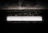 A bright fluorescent light fixture is set against a dark, textured background. The light is encased in a protective, wire mesh cover, casting shadows onto the surrounding surface.