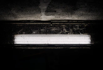 A bright fluorescent light fixture is set against a dark, textured background. The light is encased in a protective, wire mesh cover, casting shadows onto the surrounding surface.