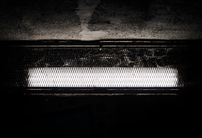 A bright fluorescent light fixture is set against a dark, textured background. The light is encased in a protective, wire mesh cover, casting shadows onto the surrounding surface.