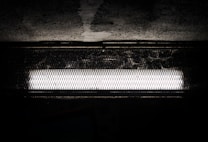 A bright fluorescent light fixture is set against a dark, textured background. The light is encased in a protective, wire mesh cover, casting shadows onto the surrounding surface.