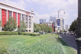 A vibrant cityscape featuring a large red and white building with the name 'KANAL' on it. The building is surrounded by lush green trees and neatly trimmed grass. In the background, modern high-rise buildings tower over the scene. A busy road runs alongside the grassy area, with vehicles and streetlights visible. A billboard with a geometric design is positioned next to the road.