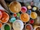 Several buckets and containers are filled with various colors of paint, including shades of orange, red, green, yellow, and black. They are placed on a rough, possibly concrete surface that appears to be part of a workshop or studio.