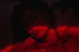 A close-up of two people in an intimate embrace illuminated by red light. The image has a dark and moody atmosphere, with one person resting their head against the other, eyes closed. Tattoos and shadows add to the mysterious vibe.