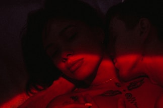 A close-up of two people in an intimate embrace illuminated by red light. The image has a dark and moody atmosphere, with one person resting their head against the other, eyes closed. Tattoos and shadows add to the mysterious vibe.