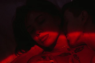 A moody, high-contrast photo of a couple silhouetted against a dark background with subtle red and blue lighting highlighting their intense connection.