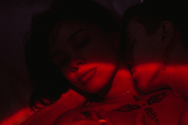 A close-up of two people in an intimate embrace illuminated by red light. The image has a dark and moody atmosphere, with one person resting their head against the other, eyes closed. Tattoos and shadows add to the mysterious vibe.