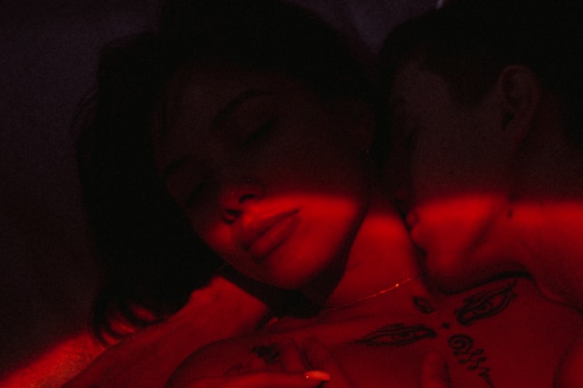 A close-up of two people in an intimate embrace illuminated by red light. The image has a dark and moody atmosphere, with one person resting their head against the other, eyes closed. Tattoos and shadows add to the mysterious vibe.