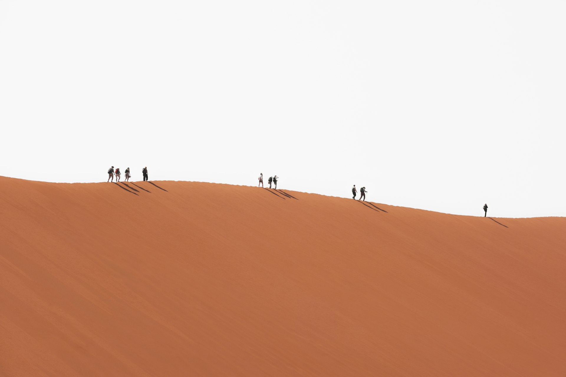 A line of small figures walking the crest of a vast red sand dune.