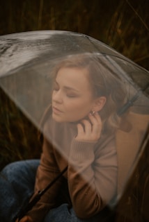 A person with closed eyes sits under a transparent umbrella, suggesting a sense of contemplation. The individual is dressed in a brown sweater and blue jeans, with an expression that conveys calmness. The background appears to be a natural setting, likely a field or meadow.
