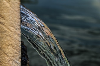 A stream of clear water flows smoothly over the side of a textured stone surface, with ripples and reflections visible. The background is a blurred body of water, suggesting a natural or serene environment.