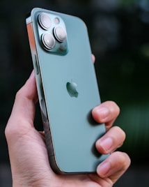 A close-up image of a hand holding a green smartphone which features three prominent camera lenses on the back. The device has a sleek, metallic finish with a brand logo in the center. The thumb and fingers are visible, indicating a firm grip on the phone.