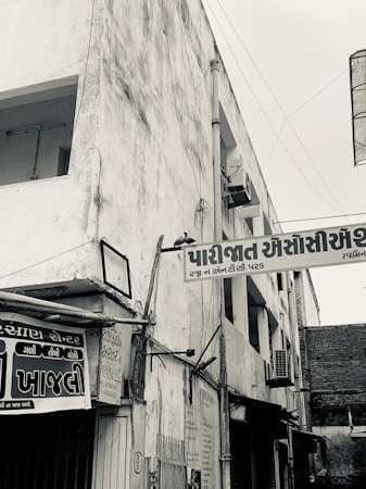 A black and white photo of an urban street corner with visible signs in an Indian script. The building appears worn with visible utility wiring. There's a sense of age and history in the architecture and surroundings.