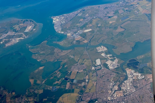 An aerial view of a coastal region showcasing a landscape with a mixture of urban and rural areas interspersed with waterways and fields. The city areas are densely packed with buildings, roads, and industrial complexes. The countryside is composed of green fields and patches of trees. The coastline features a mix of natural and developed sections, with clear separations between land and sea.