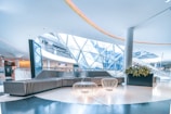 A modern indoor space with sleek, curving architectural elements. A large, contemporary seating area is positioned in the center, featuring a curved sofa and intricate wire-frame tables. Overhead, a unique geometric glass ceiling allows natural light to stream in, giving the space an open and airy feel. Nearby, a planter with vibrant green plants adds a touch of nature. The area is spacious, well-lit, and futuristic in design.