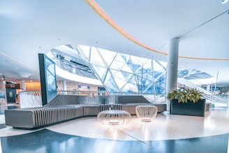 A modern indoor space with sleek, curving architectural elements. A large, contemporary seating area is positioned in the center, featuring a curved sofa and intricate wire-frame tables. Overhead, a unique geometric glass ceiling allows natural light to stream in, giving the space an open and airy feel. Nearby, a planter with vibrant green plants adds a touch of nature. The area is spacious, well-lit, and futuristic in design.