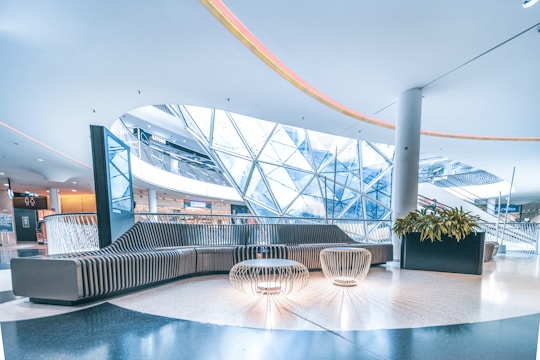 A modern indoor space with sleek, curving architectural elements. A large, contemporary seating area is positioned in the center, featuring a curved sofa and intricate wire-frame tables. Overhead, a unique geometric glass ceiling allows natural light to stream in, giving the space an open and airy feel. Nearby, a planter with vibrant green plants adds a touch of nature. The area is spacious, well-lit, and futuristic in design.