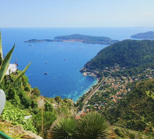 A coastal landscape featuring a vibrant blue sea dotted with several small boats. In the foreground, there are lush green plants and rugged cliffs leading down to a winding road that runs parallel to the shoreline. The coastal area is lined with trees and small houses scattered throughout the hills.