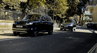 Two black SUVs are driving along a street lined with trees and buildings. Metallic barriers separate the road from the pedestrian area. The sky is overcast, casting shadows on the road.