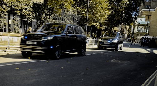 Two black SUVs are driving along a street lined with trees and buildings. Metallic barriers separate the road from the pedestrian area. The sky is overcast, casting shadows on the road.