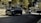 Two black SUVs are driving along a street lined with trees and buildings. Metallic barriers separate the road from the pedestrian area. The sky is overcast, casting shadows on the road.
