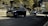 Two black SUVs are driving along a street lined with trees and buildings. Metallic barriers separate the road from the pedestrian area. The sky is overcast, casting shadows on the road.