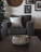 A cozy living room setting with a focus on a textured, rustic bowl on a dark wooden table. In the background, a plush armchair is adorned with a soft white pillow. A large clock and several framed pictures decorate the wall, adding to the warm, homey atmosphere.