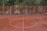 A wet, deserted sports court with an orange-red surface is shown with a white half-circle line marking in the center. Surrounding the court is a metal fence painted in orange. Behind the fence, there are trees with green foliage and buildings partially visible in the background. A few cars are parked along the street.