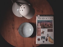 A wooden table holds a travel guidebook titled 'Morocco', featuring visuals of cultural landmarks. Beside it is a white bowl with a geometric pattern and a lamp with a perforated shade.