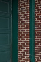 A section of a building features a red brick wall with green paint accents, including a door frame and gutter. The bricks are arranged in a horizontal pattern, and the green elements provide stark contrast against the earthy brick tones.