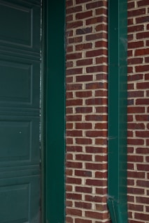 A section of a building features a red brick wall with green paint accents, including a door frame and gutter. The bricks are arranged in a horizontal pattern, and the green elements provide stark contrast against the earthy brick tones.