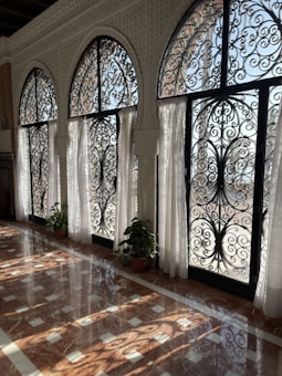 Intricately designed wrought iron windows with arched tops, adorned with floral patterns, allow soft natural light to fill the space. White curtains add elegance, and potted plants at the base of each window bring a touch of greenery. The polished marble floor reflects the complex patterns of sunlight and shadow.