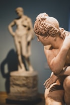 A close-up view of a classical marble statue, capturing intricate details like the curls in the hair and the contours of the body. In the background, another blurred statue stands, creating depth and contrast. The lighting casts soft shadows, highlighting the smooth texture of the marble.