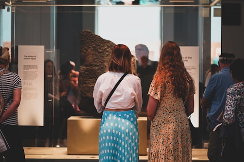 Several people are gathered around a display case in a museum. Two women are prominently seen from behind, observing a large, stone artifact inside the case. The artifact appears to be an ancient stone with inscriptions. Informational plaques are visible beside the display, presumably providing context about the exhibit.