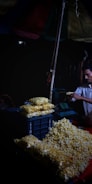 A street vendor is preparing and packaging popcorn under a canopy. The scene is dimly lit, with heaps of popcorn piled on a table and packed into plastic bags. The vendor appears focused on his task.