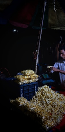 A street vendor is preparing and packaging popcorn under a canopy. The scene is dimly lit, with heaps of popcorn piled on a table and packed into plastic bags. The vendor appears focused on his task.