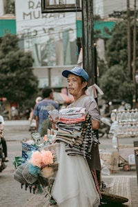 A street vendor stands with a variety of items for sale, including colorful towels, sponges, and other household goods. The vendor is wearing a blue cap and a gray shirt, and the scene is set in a bustling urban environment with people and blurred background details.