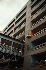 A multi-story parking structure with visible concrete surfaces and white railings surrounds the building. Each section is highlighted with a different vibrant color, including orange, green, and blue. There's a covered pedestrian bridge connecting the floors with a row of ceiling lights. A lamp is suspended from a wire in the foreground, and a no-entry traffic sign is at the bottom right.