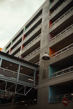 A multi-story parking structure with visible concrete surfaces and white railings surrounds the building. Each section is highlighted with a different vibrant color, including orange, green, and blue. There's a covered pedestrian bridge connecting the floors with a row of ceiling lights. A lamp is suspended from a wire in the foreground, and a no-entry traffic sign is at the bottom right.