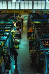 Inside a dimly lit market hall, various stalls display an assortment of fruits, vegetables, and groceries. A few people are browsing and tending to their items. The atmosphere feels calm yet bustling with a variety of produce including bananas, oranges, and packaged goods are visible.
