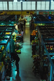 Inside a dimly lit market hall, various stalls display an assortment of fruits, vegetables, and groceries. A few people are browsing and tending to their items. The atmosphere feels calm yet bustling with a variety of produce including bananas, oranges, and packaged goods are visible.