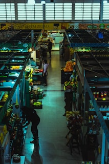 Inside a dimly lit market hall, various stalls display an assortment of fruits, vegetables, and groceries. A few people are browsing and tending to their items. The atmosphere feels calm yet bustling with a variety of produce including bananas, oranges, and packaged goods are visible.