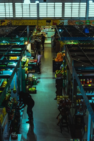 Inside a dimly lit market hall, various stalls display an assortment of fruits, vegetables, and groceries. A few people are browsing and tending to their items. The atmosphere feels calm yet bustling with a variety of produce including bananas, oranges, and packaged goods are visible.