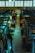 Inside a dimly lit market hall, various stalls display an assortment of fruits, vegetables, and groceries. A few people are browsing and tending to their items. The atmosphere feels calm yet bustling with a variety of produce including bananas, oranges, and packaged goods are visible.