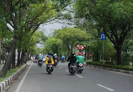A group of people riding motorcycles down a tree-lined street. The road is flanked by lush green trees and there are several motorcycles visible, with riders wearing helmets and various colored jackets. A blue traffic sign indicating a U-turn is visible on the right side.