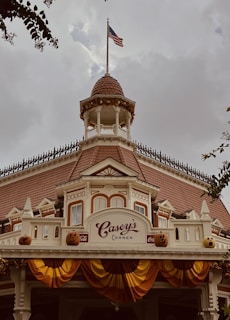 A vintage-style building with detailed architectural elements like a turret and ornamental trim. The American flag flies at the top, and festive decorations including carved pumpkins and autumnal garlands adorn the facade. The sign reads 'Casey's Corner' with Coca-Cola logos.