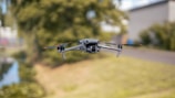 A drone hovers in mid-air against a blurred outdoor background with trees and a building. The drone is small, with a camera mounted on its front, and four propellers visible.
